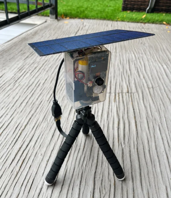 The BalconyPatrol device on a balcony deck — solar panel on top, transparent enclosure with the camera and electronics visible, mounted on a flexible tripod