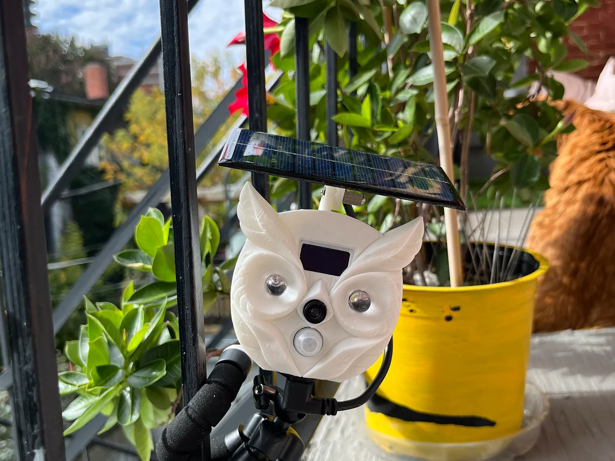The owl-shaped BalconyPatrol prototype on a balcony railing, a ginger cat sitting behind it among the plants